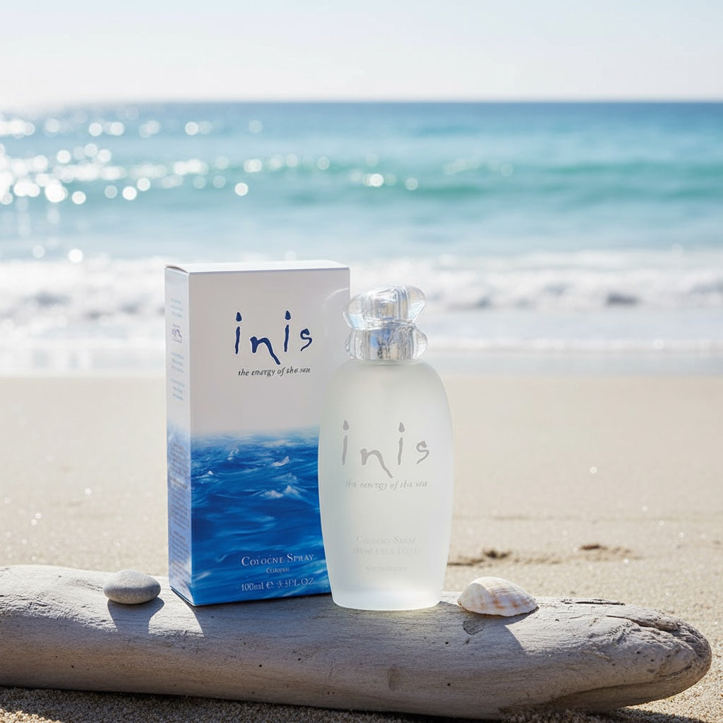 Inis Cologne Spray 100ml bottle and box on driftwood on a sandy beach with ocean waves in background
