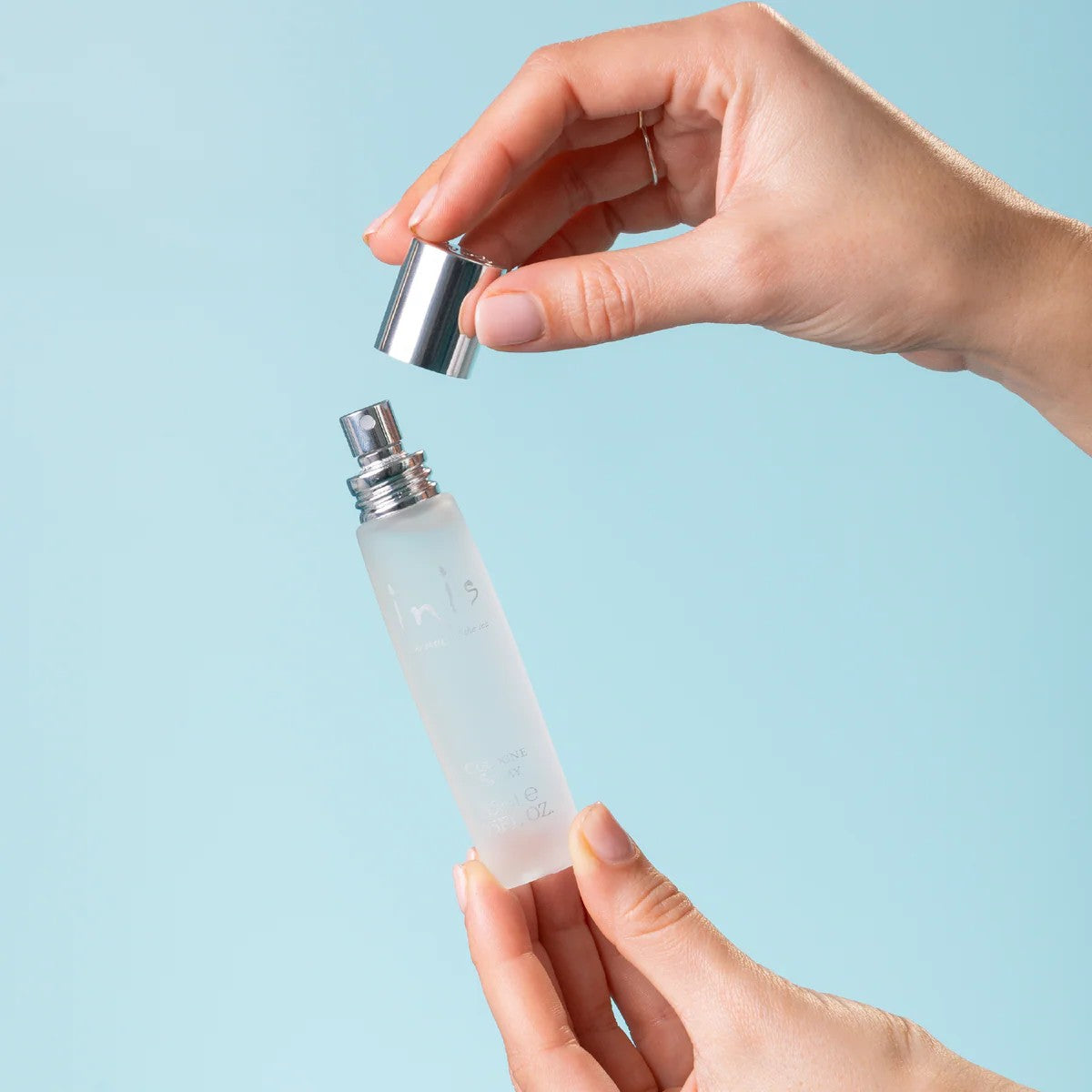 Hands removing the silver cap from an Inis Cologne Spray 15ml bottle on a blue background