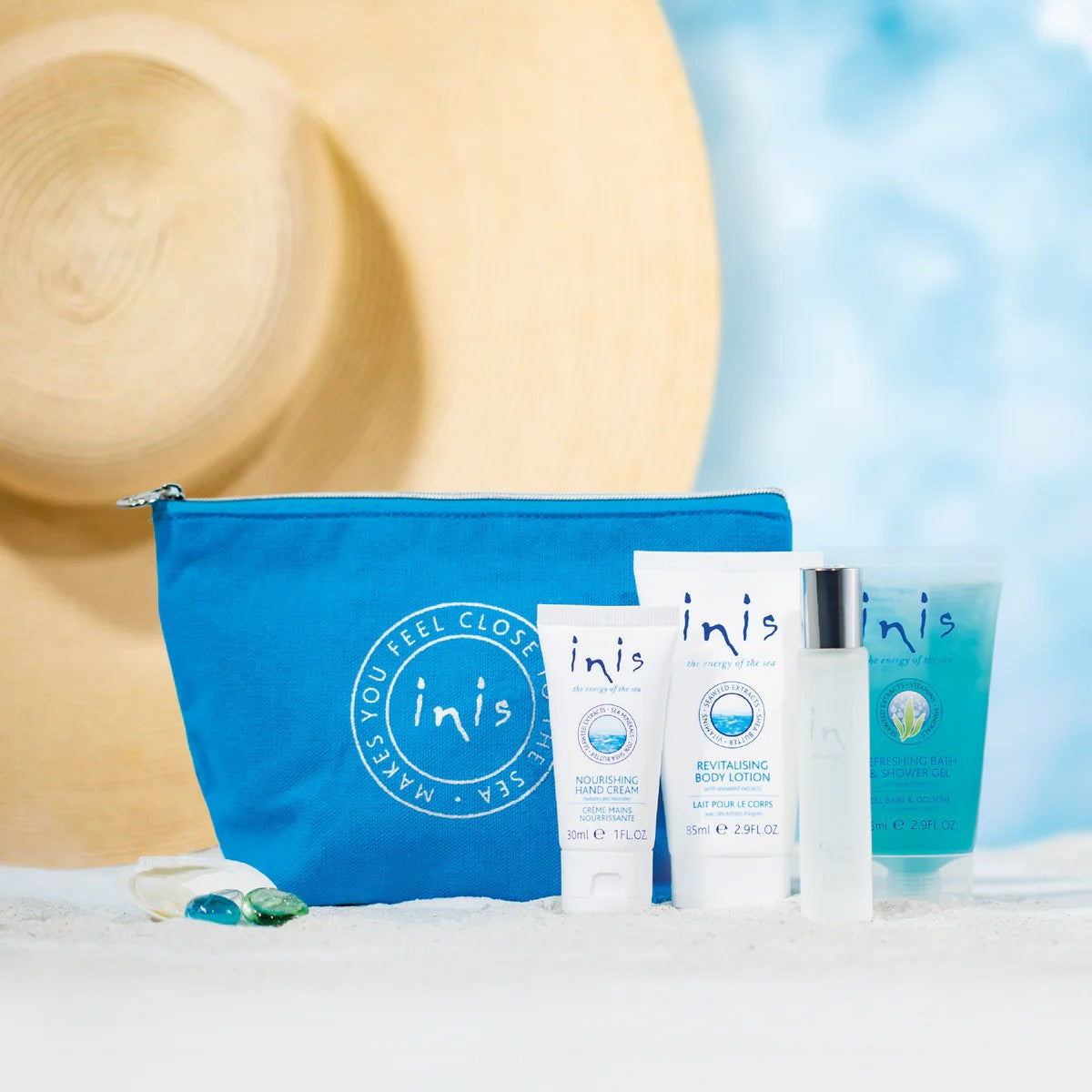 Inis Voyager Set products and blue zip bag arranged on sand in front of a sun hat with a beach sky background